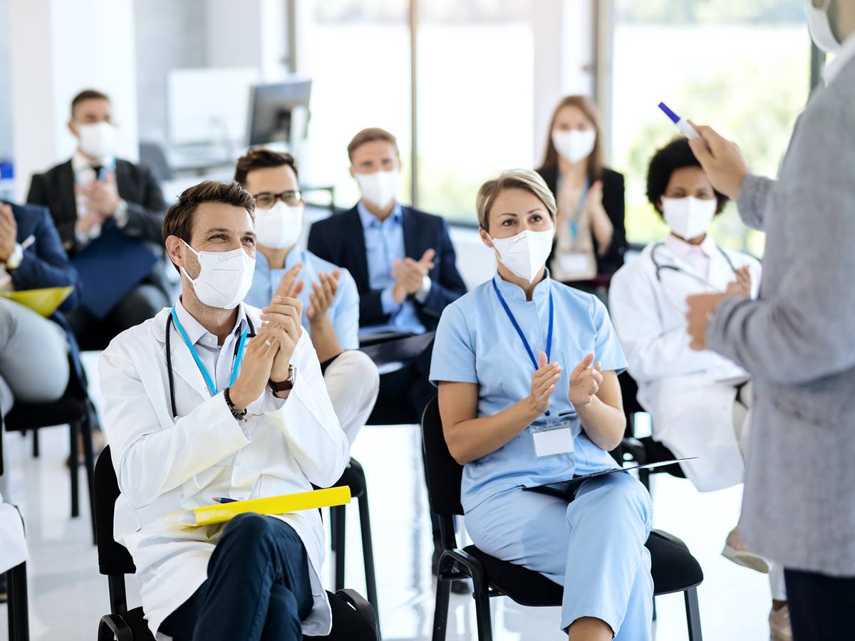 Healthcare workers and business people wearing face masks and ap Large group of doctors and business people applauding while attending educational event during coronavirus pandemic. Focus is on male doctor.