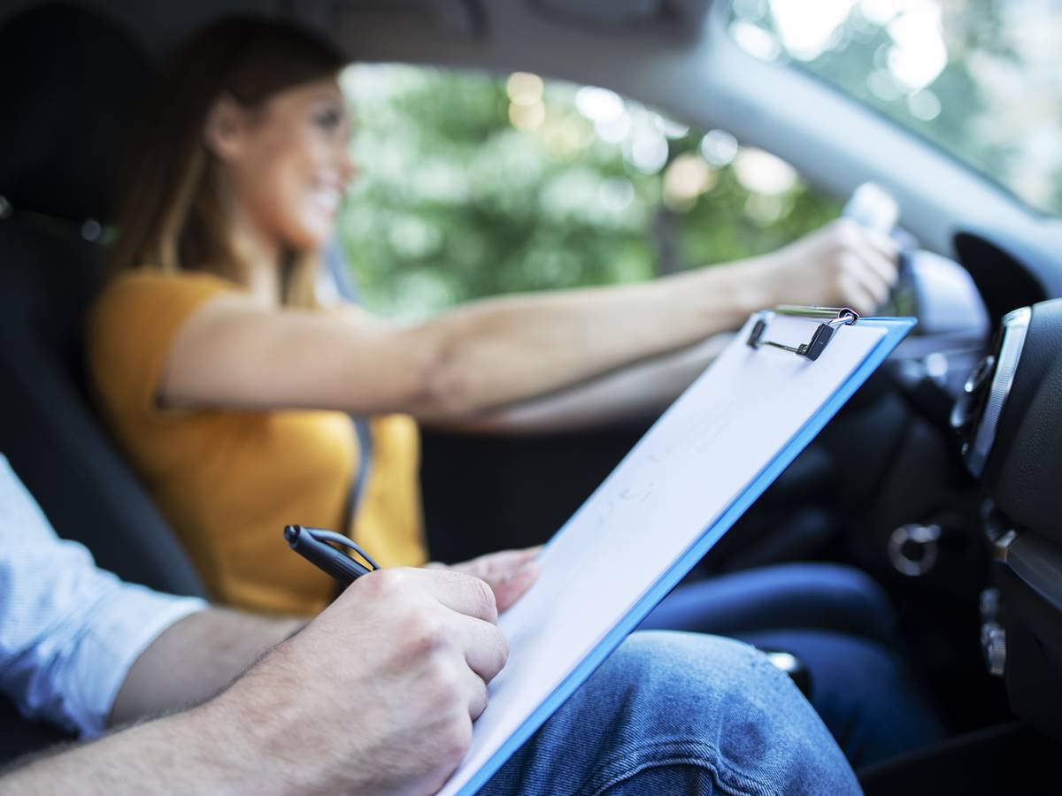 Close up view of driving instructor holding checklist while in background female student steering and driving car.