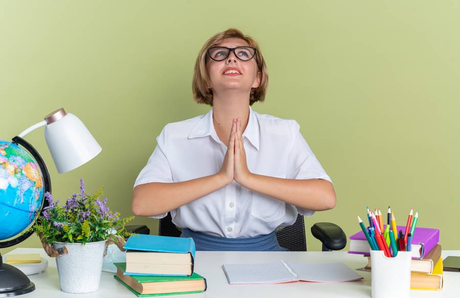anxious young blonde student girl wearing glasses sitting at desk with school tools looking up keeping hands together praying isolated on olive green background