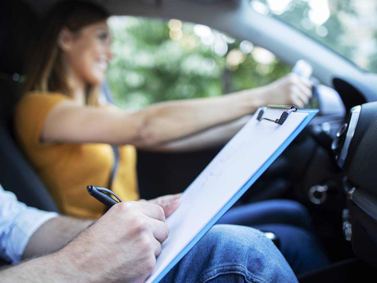 Close up view of driving instructor holding checklist while in background female student steering and driving car.