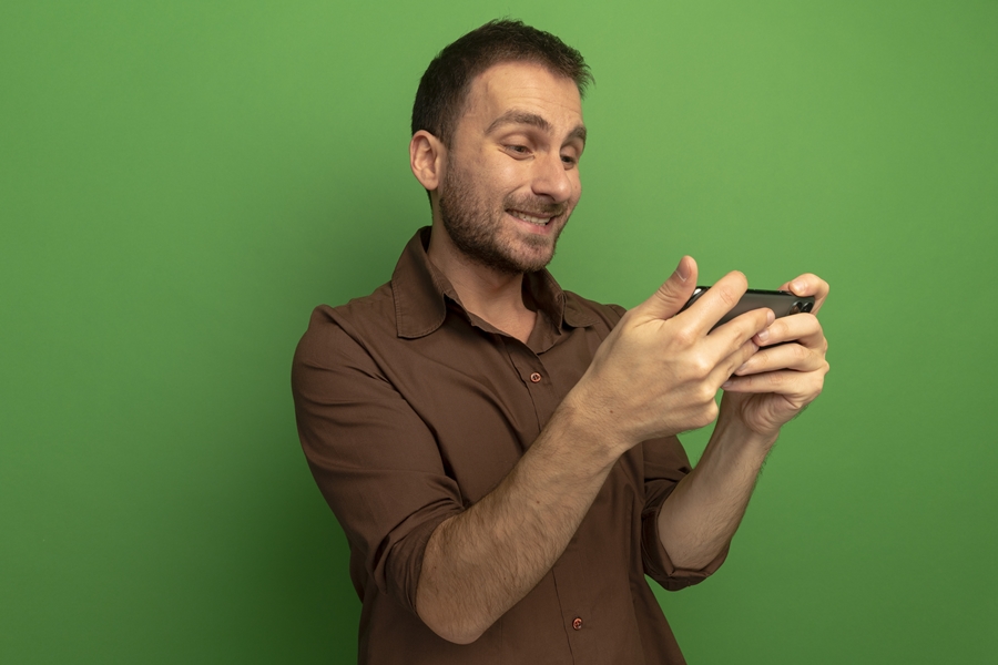 smiling young caucasian man holding and looking at mobile phone isolated on green background with copy space smiling young caucasian man holding and looking at mobile phone isolated on green background with copy space