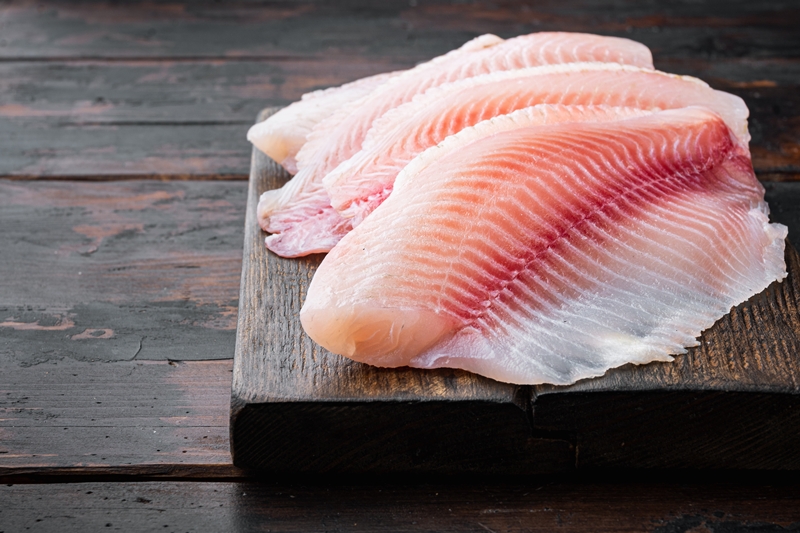 Raw tilapia fish fillet meat cuts, on dark wooden background
