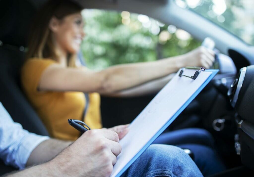Close up view of driving instructor holding checklist while in background female student steering and driving car.