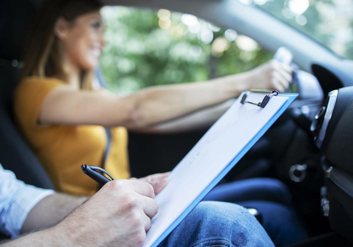 Close up view of driving instructor holding checklist while in background female student steering and driving car.