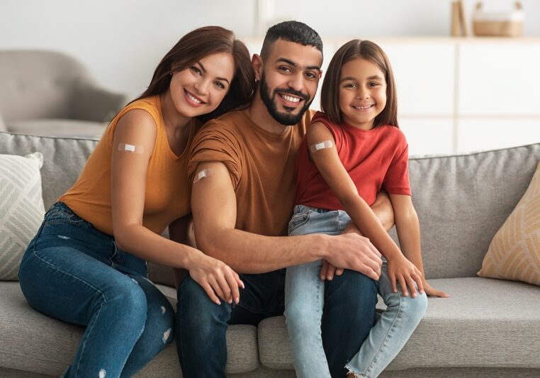 Coronavirus Vaccination Advert. Happy Vaccinated Family Of Three People Showing Arm With Sticking Plaster After Covid-19 Vaccine Vax Injection Posing Sitting On Couch In Living Room, Smiling To Camera