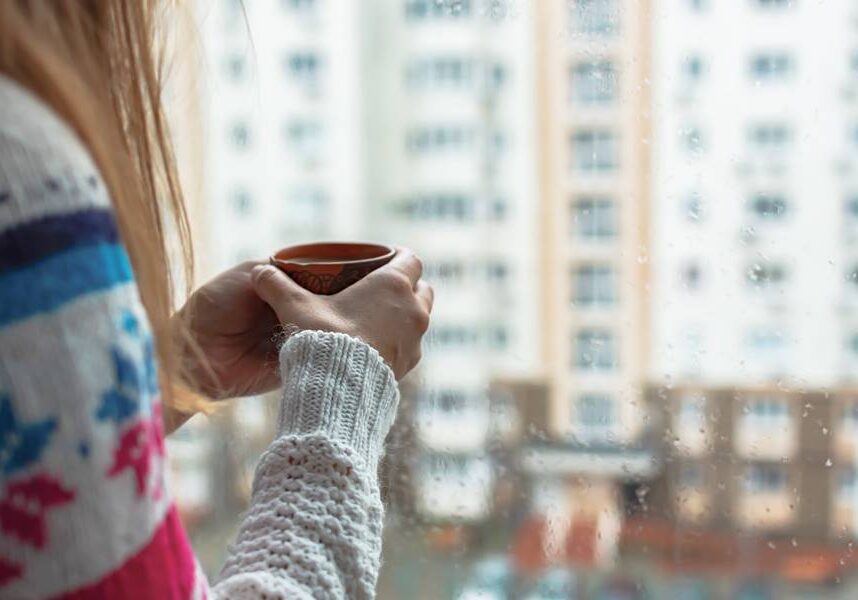 A girl with a mug in her hands at the window. Morning coffee on a rainy day. The concept of autumn sadness, cold and not cozy mood. It's raining outside. Warm house romance comfort. Copy space