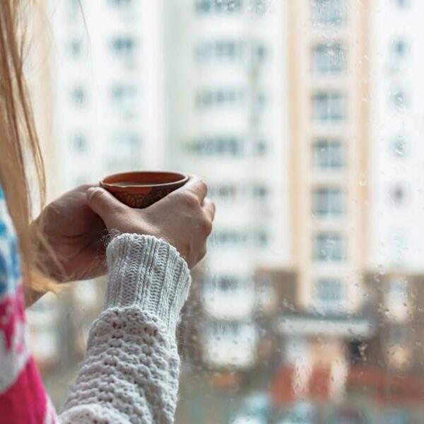 A girl with a mug in her hands at the window. Morning coffee on a rainy day. The concept of autumn sadness, cold and not cozy mood. It's raining outside. Warm house romance comfort. Copy space