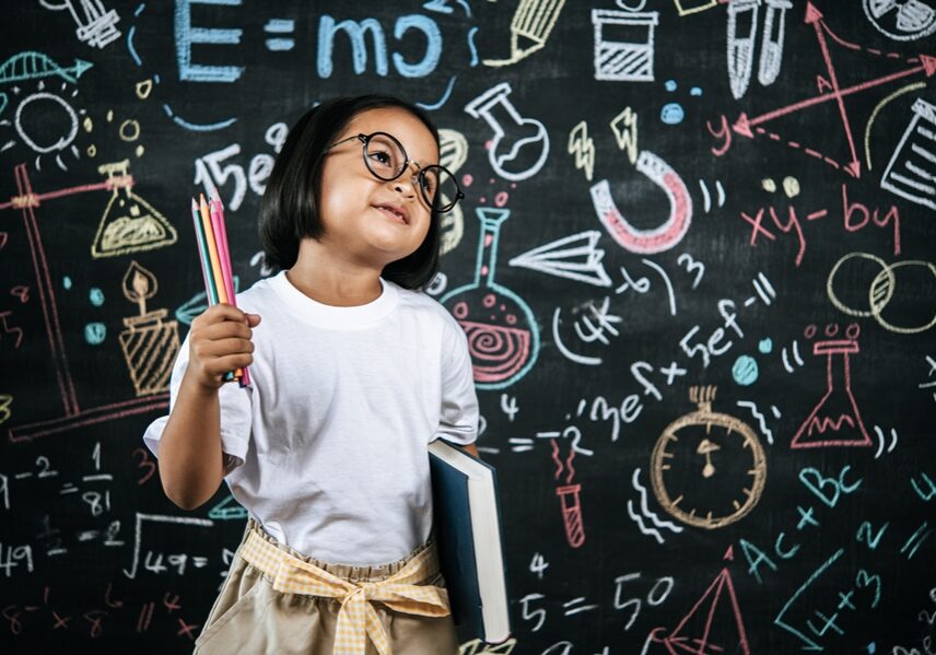 Selective focus, Little girl wearing eyeglasses holding colors pencil in hand and holding large textbook standing at front of blur blackboard in background, smile with happy