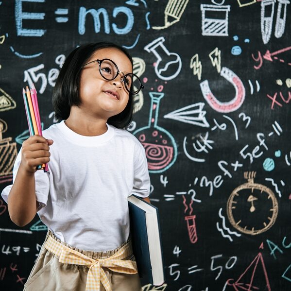 Selective focus, Little girl wearing eyeglasses holding colors pencil in hand and holding large textbook standing at front of blur blackboard in background, smile with happy