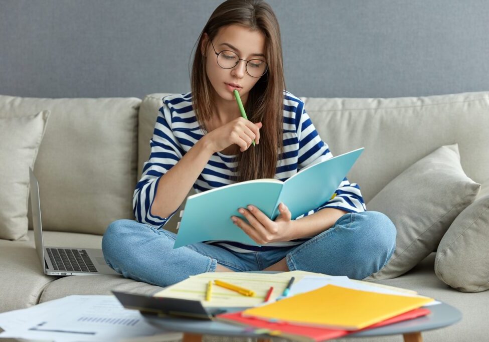 Studying online concept. Serious young woman being busy with remote freelance project, sits at comfortable sofa, writes notes, holds textbook, use laptop computer at home with wireless internet