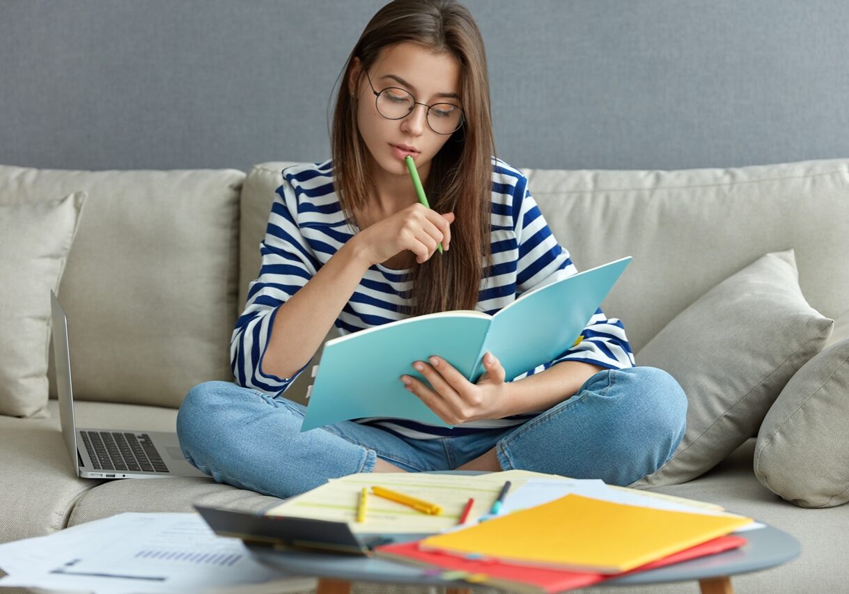 Studying online concept. Serious young woman being busy with remote freelance project, sits at comfortable sofa, writes notes, holds textbook, use laptop computer at home with wireless internet